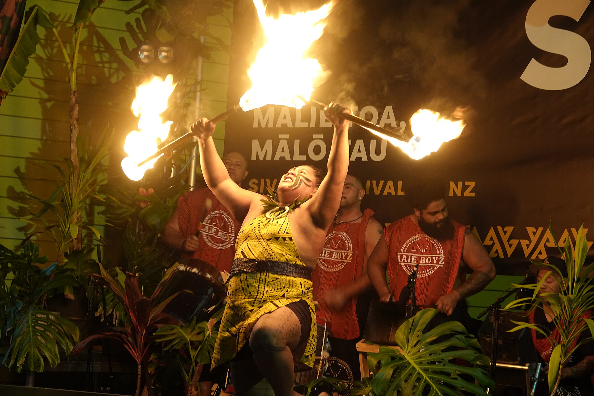 A Samoan fire-knife dancer performing on stage with drummers behind, at the Siva Afi festival in Māngere, New Zealand, 2024