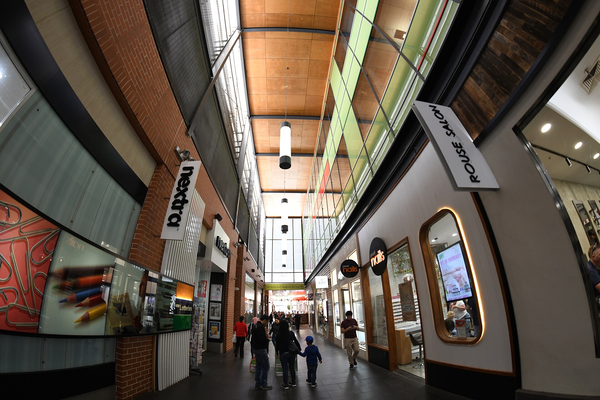 A narrow covered walkway at Rouse Hill Town Centre with a newsagency, nail salon and families walking